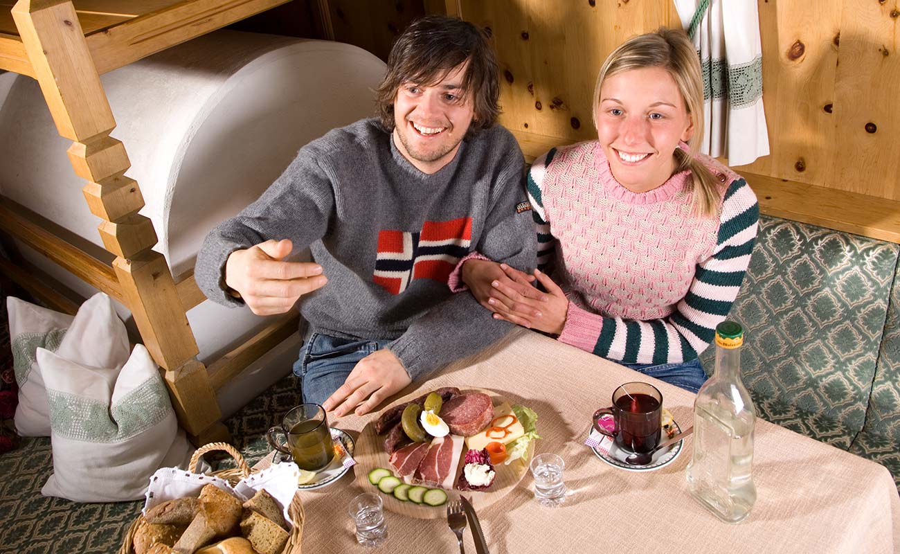 Pärchen am Tisch auf der Ofenbank mit Brettljause und Getränk